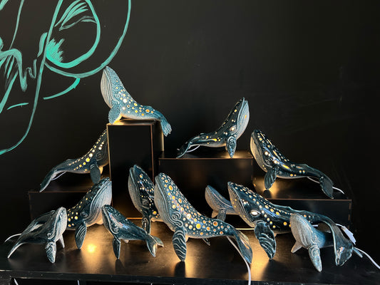 Display of handmade ceramic humpback whale lamps with glowing perforated details on black podiums against a dark background