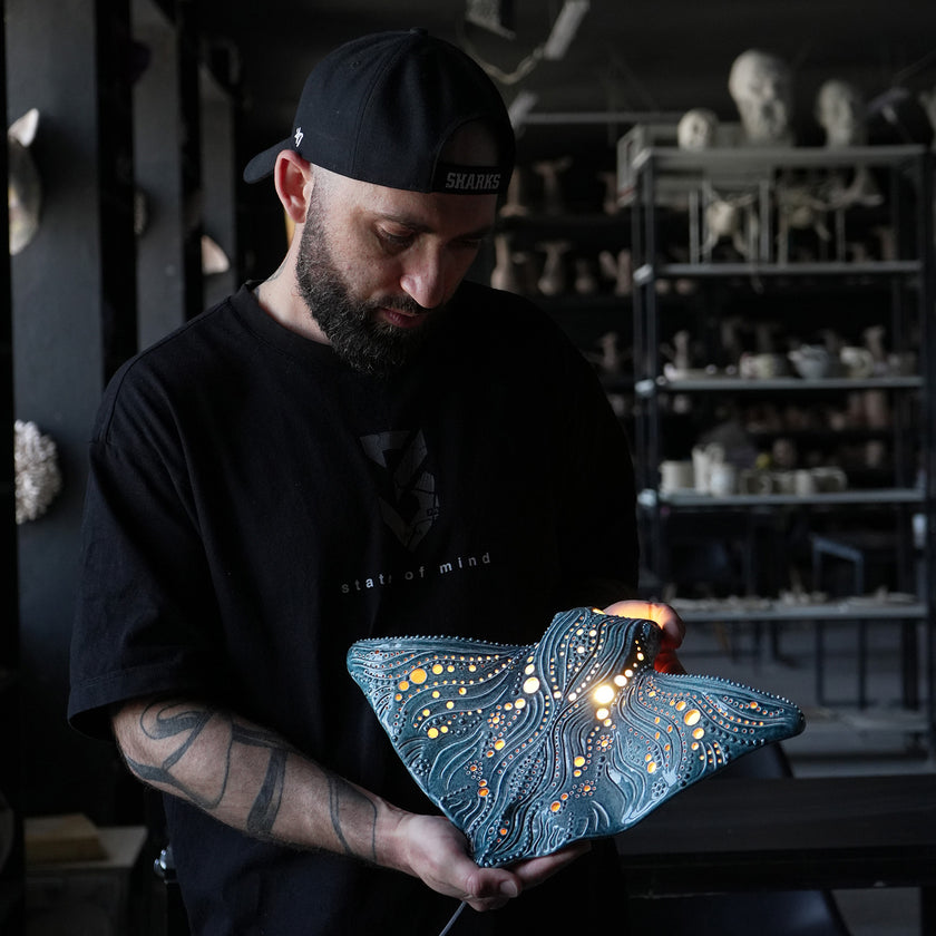 Viktor Kushchenko holding an illuminated blue ceramic manta ray wall lamp with wave patterns in the KUST studio.