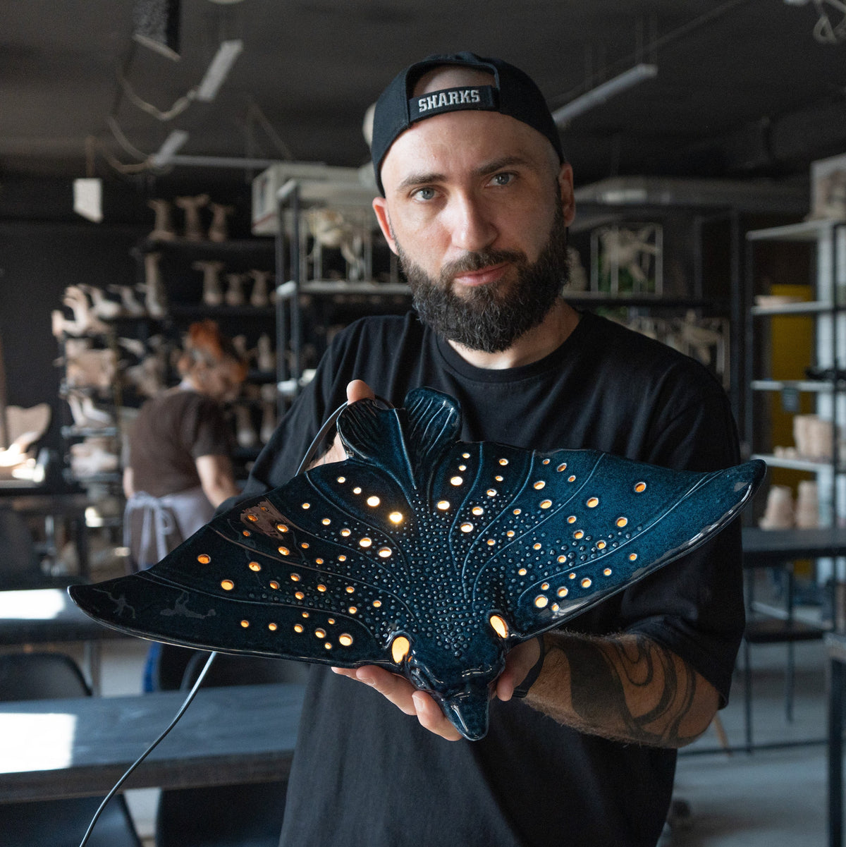 A man wearing a black cap and black t-shirt holds a dark blue manta ray-shaped decorative lamp with glowing warm lights inside what appears to be a workshop or studio.