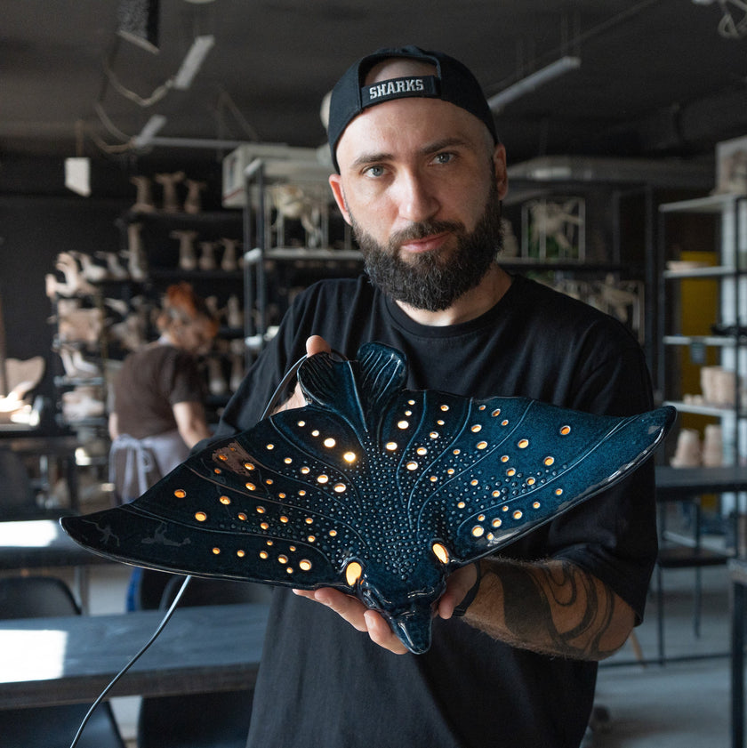 A man wearing a black cap and black t-shirt holds a dark blue manta ray-shaped decorative lamp with glowing warm lights inside what appears to be a workshop or studio.