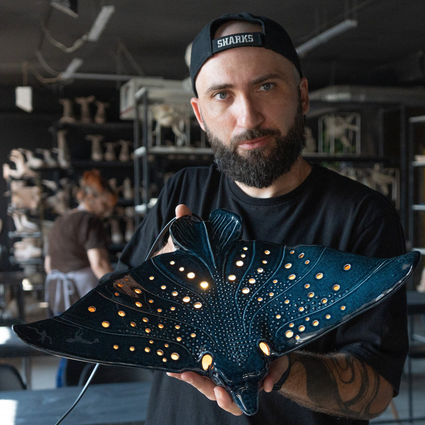 A man wearing a black t-shirt and a black cap with 'SHARKS' on it holds a large, dark blue manta ray-shaped decorative light with glowing warm yellow lights inside, set against a workshop background.