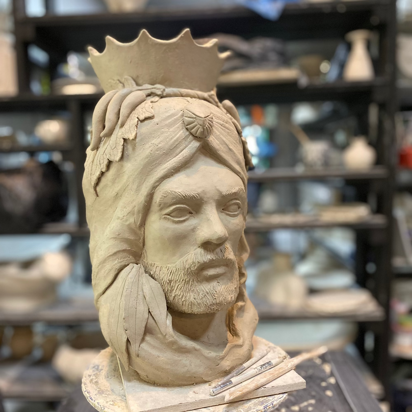 Clay sculpture of a figure with a crown on a stand in a pottery studio.